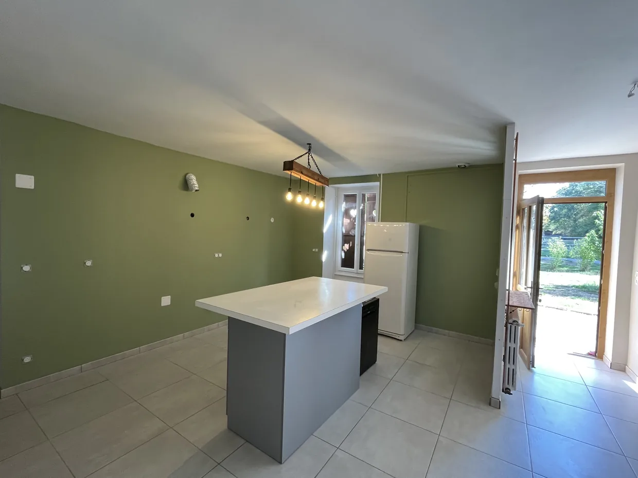La même cuisine après travaux, murs vert olive et plafond blanc, îlot central et ouverture sur le jardin