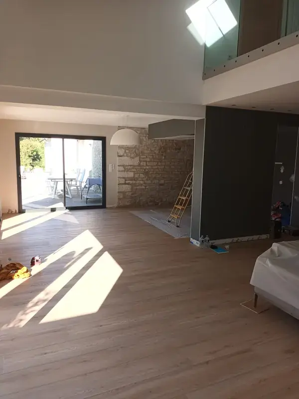 Grand volume avec mezzanine et mur de pierre, sol bois clair traversé par la lumière rasante d'une baie vitrée